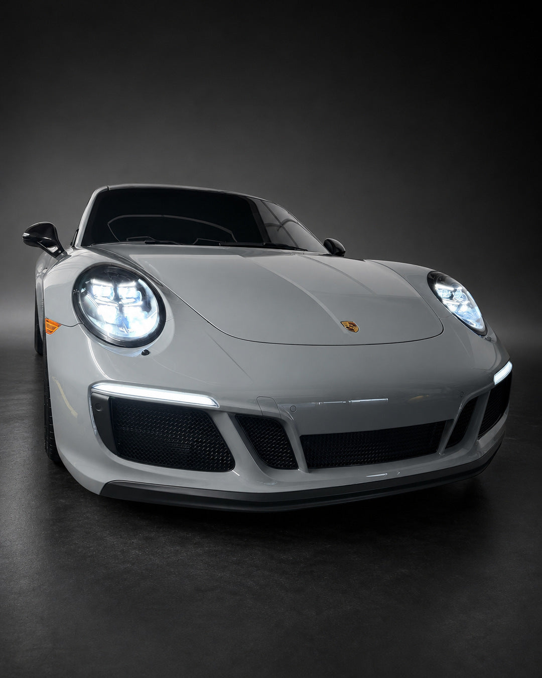 Studio-style front view of a gray Porsche 911 991.2 GTS with the V2 headlights and white DRLs illuminated