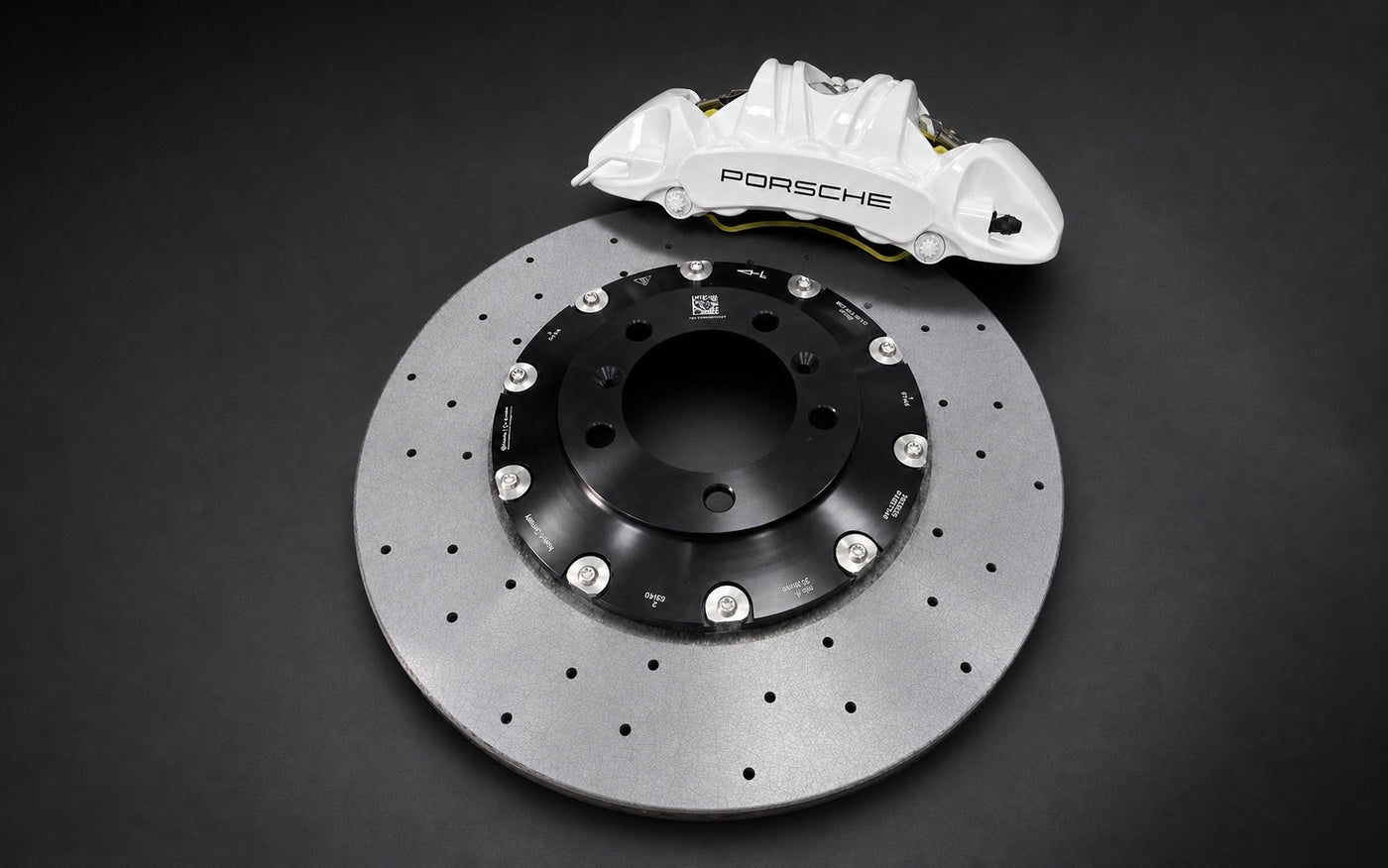 White Porsche six-piston caliper and drilled carbon-ceramic rotor photographed on a dark studio surface